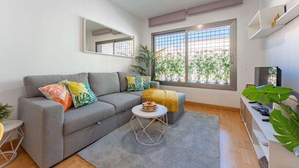Ein modernes Wohnzimmer mit grauem Sofa, bunten Kissen, einem kleinen runden Tisch mit einer Snackplatte, einem großen Fenster mit Blick ins Grüne und einem wandmontierten Fernseher.