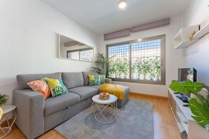Ein modernes Wohnzimmer mit grauem Sofa, bunten Kissen, einem kleinen runden Tisch mit einer Snackplatte, einem großen Fenster mit Blick ins Grüne und einem wandmontierten Fernseher.