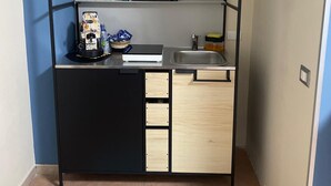 Une petite cuisine compacte avec un meuble noir et en bois clair, un petit évier et un plan de travail sur lequel se trouvent une machine à café et quelques objets décoratifs.