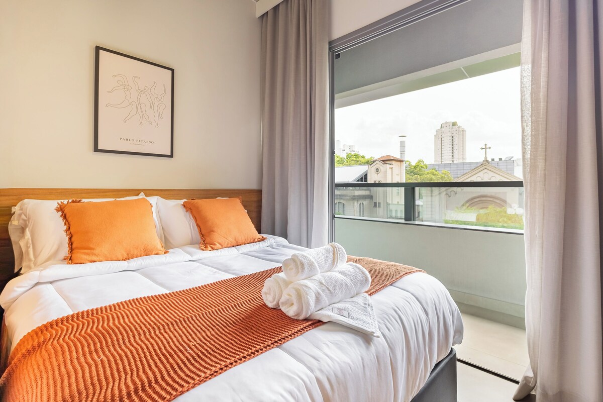 Quarto de hotel com uma cama, uma obra de arte emoldurada, uma janela com vista para a cidade e uma varanda.