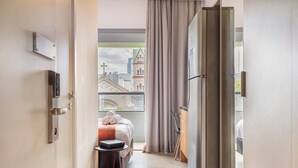 A hotel room with a bed, a table with a device, a chair, a wardrobe, and a view of a building through a window. - Piscina Gym & Rooftop no Jardins - Luxo (São Paulo)