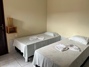 Habitación con dos camas, cada una con una colcha blanca y una toalla doblada. Se observa una repisa de madera con una cesta tejida en la pared.