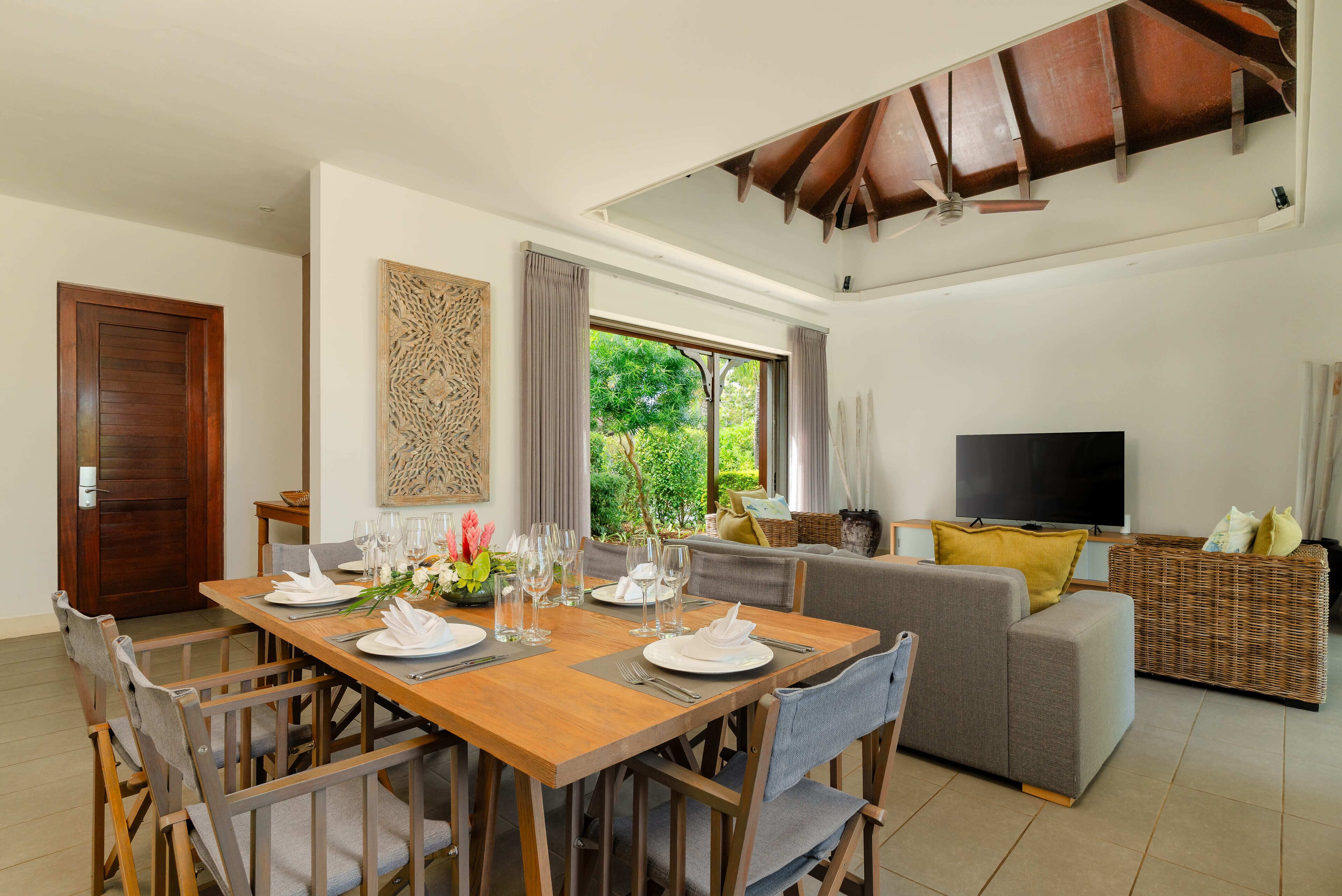 A dining area with a wooden table set for four, a sofa, a television, and a window with a view of greenery.