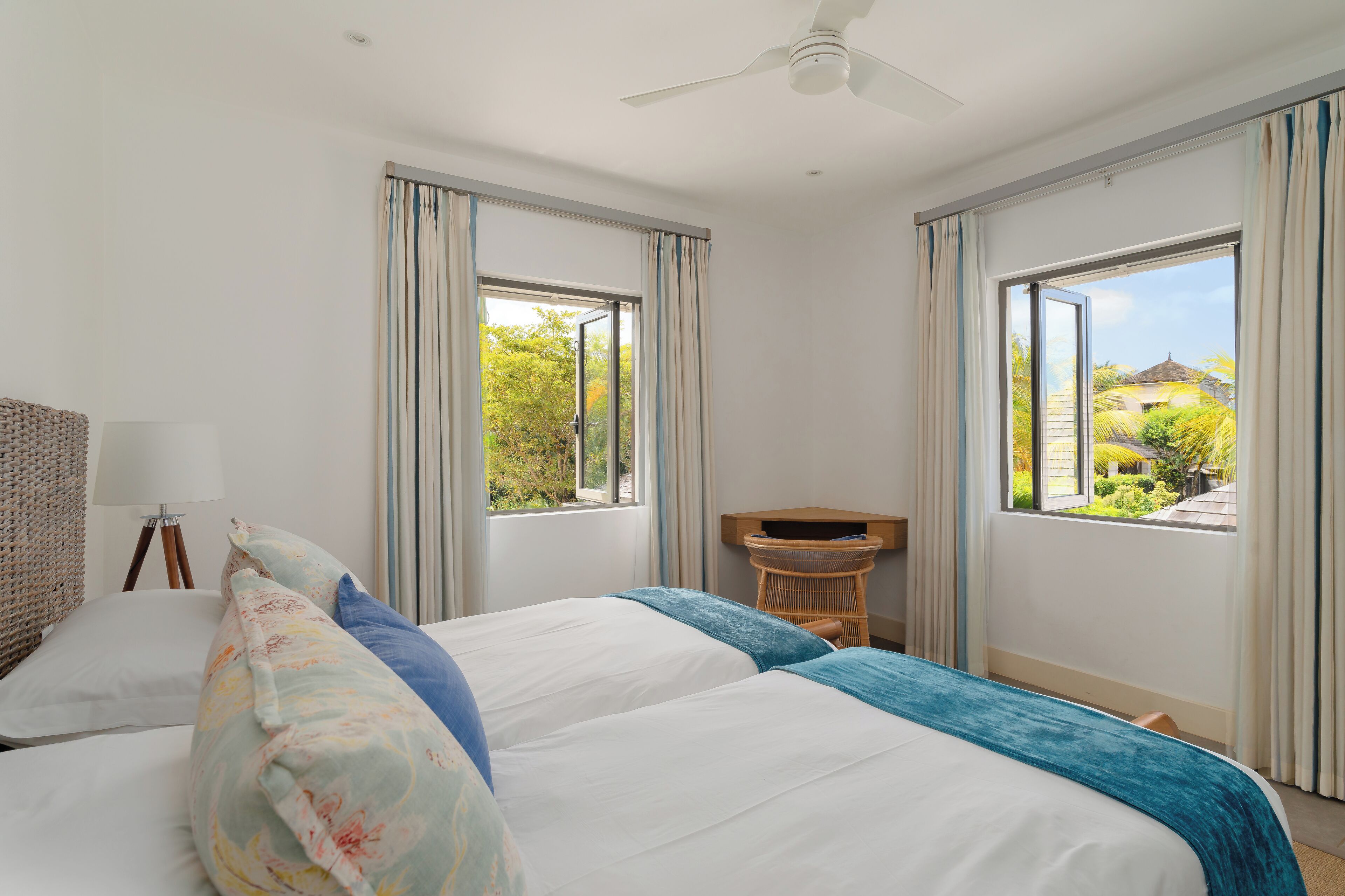 Habitación de hotel con dos camas, ventilador de techo, escritorio y ventanas amplias con cortinas.