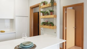 A modern kitchen with white cabinets, a white countertop, and a wooden door with a rope shelf holding potted plants. - Edifício Vencedor (Matosinhos)