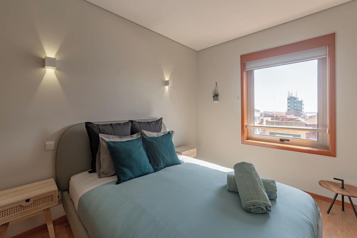 Une chambre à coucher comprenant un lit, des oreillers, une table de chevet, une petite table, une fenêtre avec vue et un luminaire fixé au mur.
