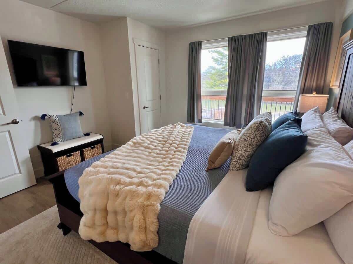 Um quarto com cama, televisão, janela com cortinas e porta.