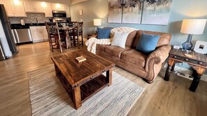 Una sala de estar con una mesa de centro de madera, un sofá con cojines azules, un comedor con sillas y una cocina al fondo.