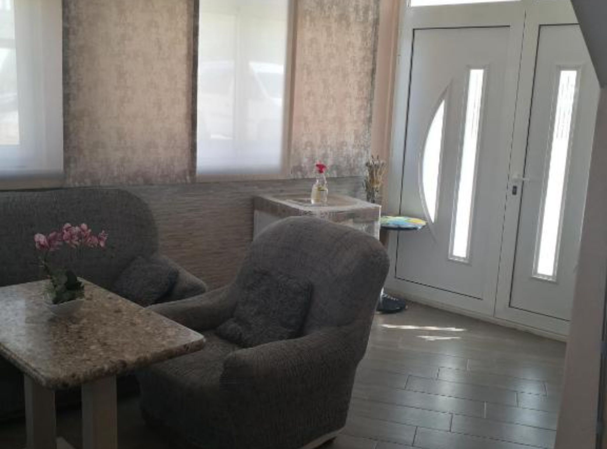 A room with a grey armchair, a small table with a vase, a window with blinds, and a door with a frosted glass panel.