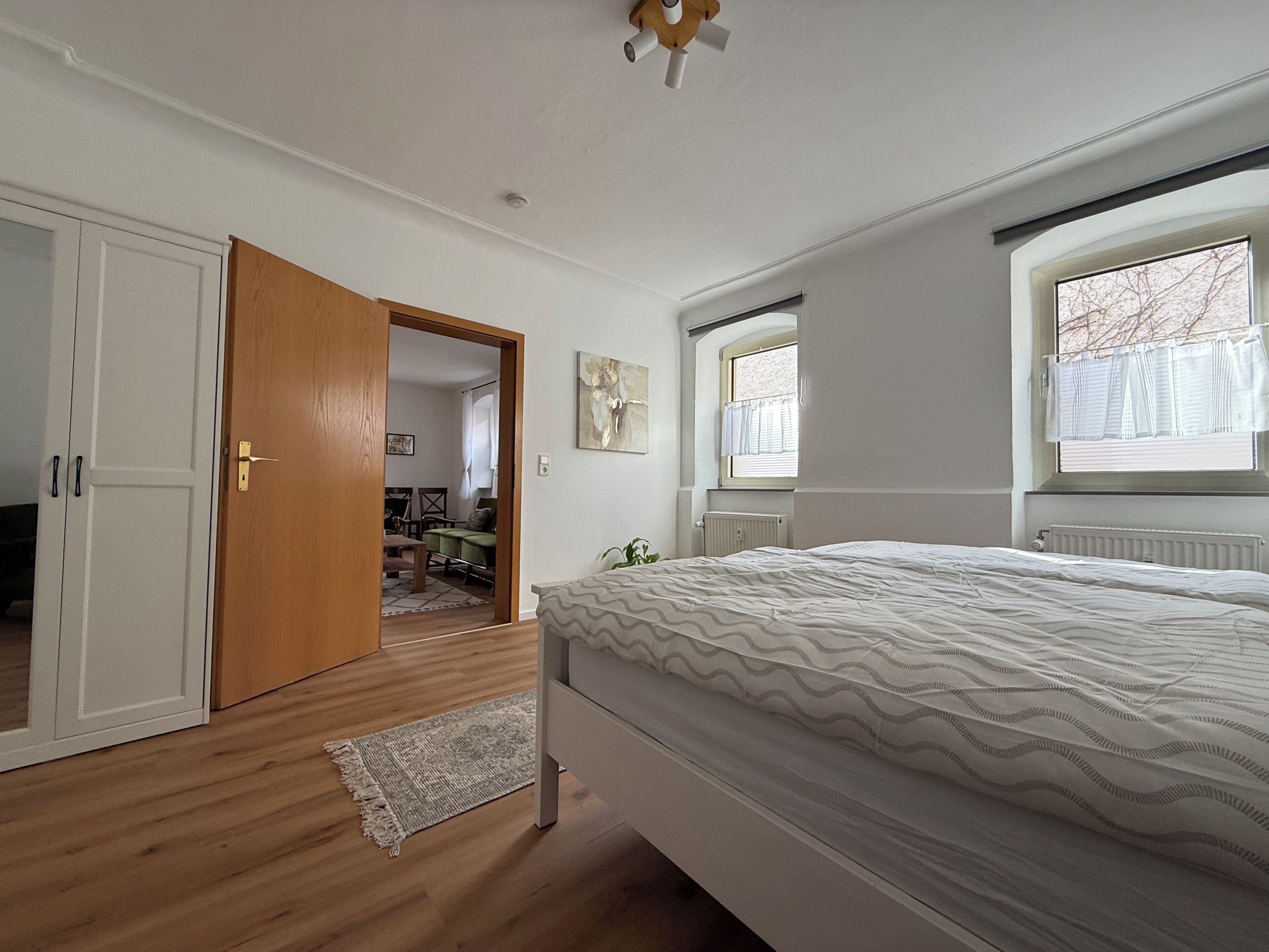 Een slaapkamer met een bed, een raam, een radiator en een deur naar een andere kamer.