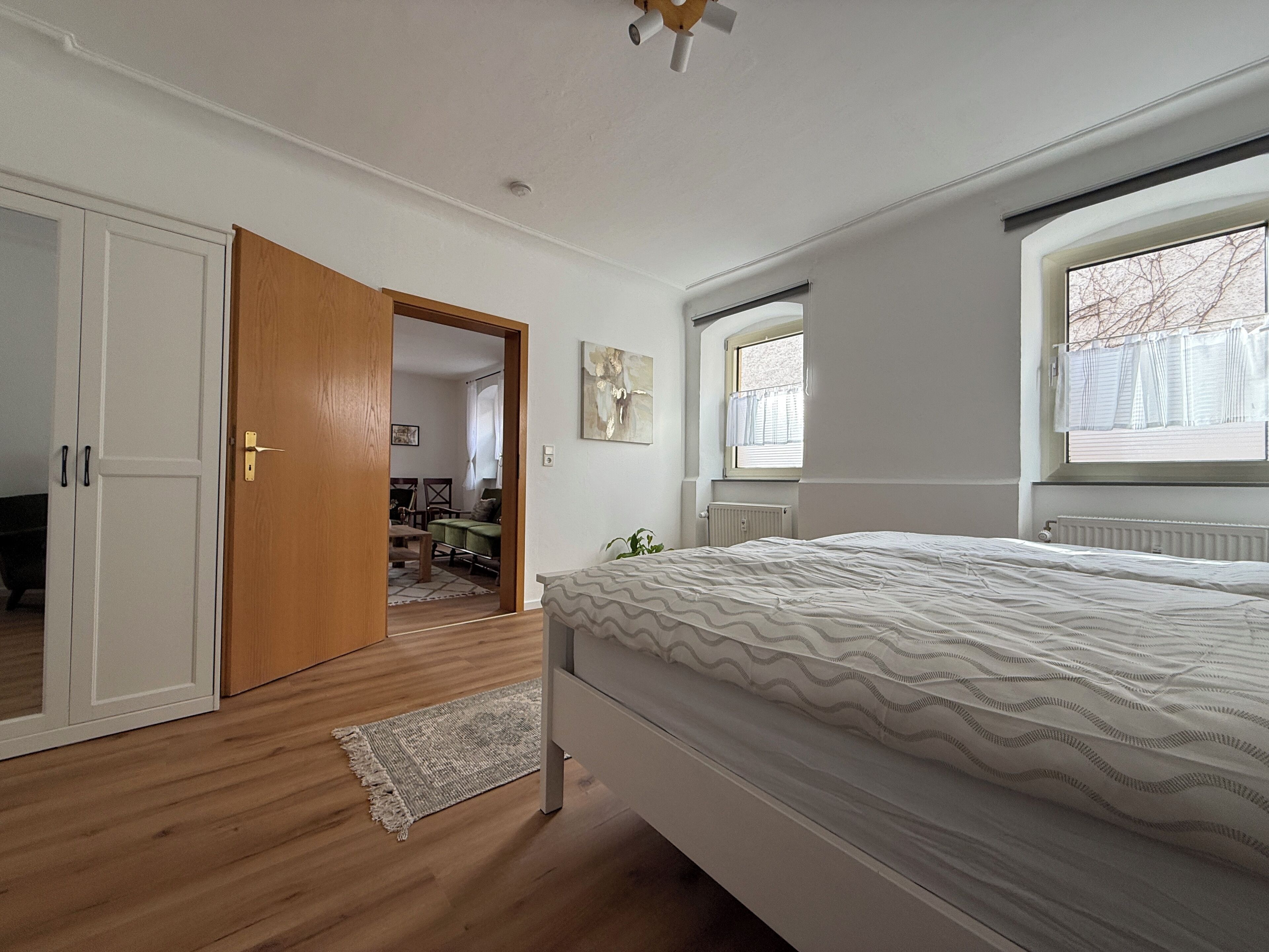 Ein Schlafzimmer mit Bett, Fenster, Heizkörper und einer Tür zu einem anderen Raum.