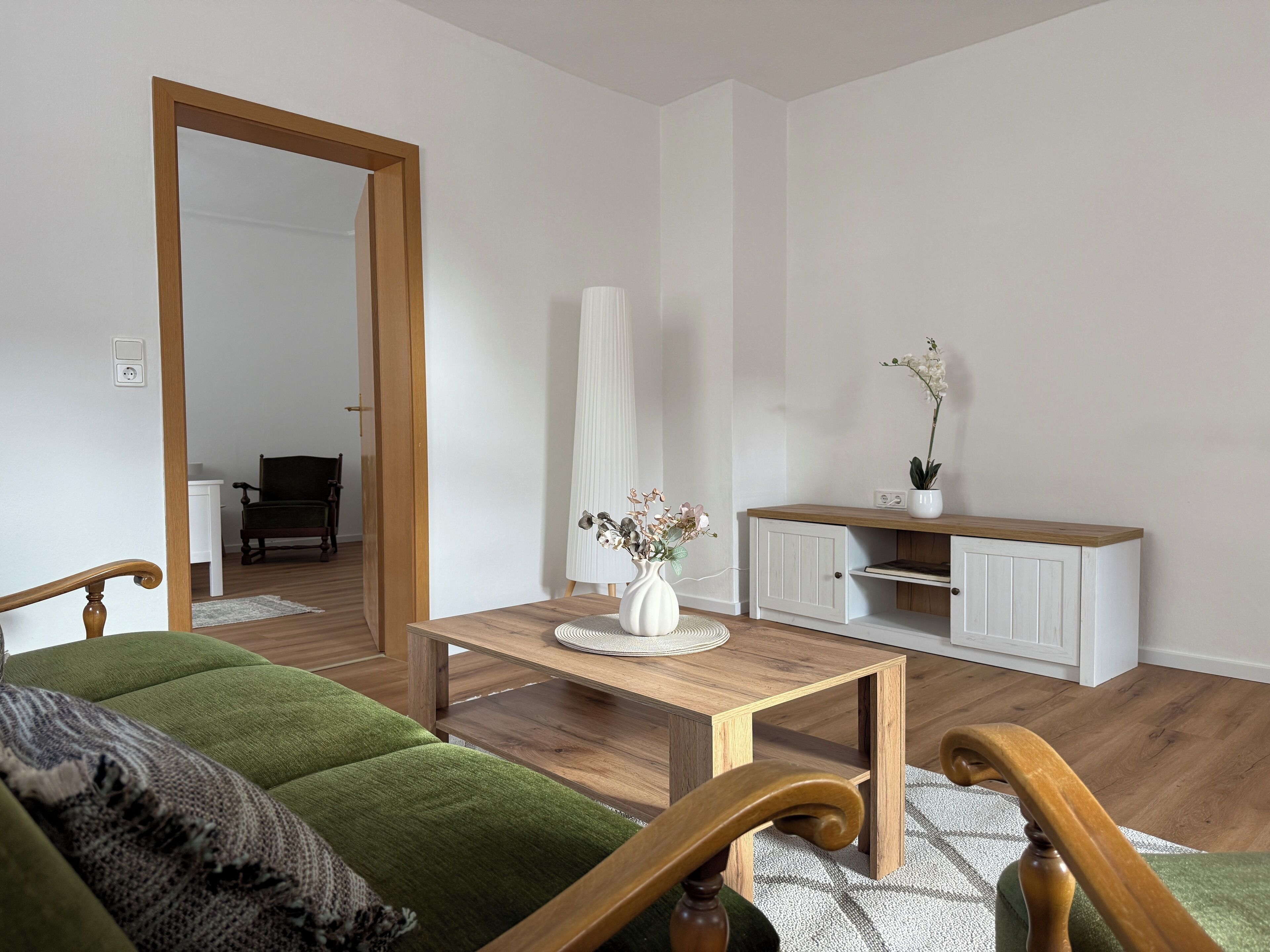 Ein Wohnzimmer mit einem grünen Sofa, einem Holztisch, einem weißen Schrank und einer Tür zu einem anderen Raum.