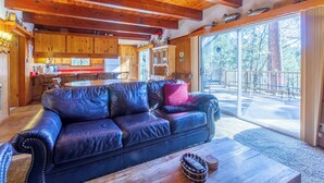 Interior acogedor de una cabaña con un sofá de cuero, vigas de techo de madera, una zona de cocina y un amplio ventanal corredero que da a una terraza exterior.
