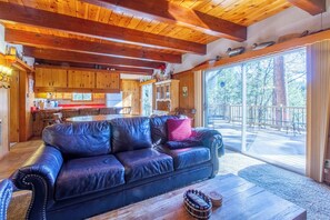Ein gemütliches Blockhaus-Interieur mit Ledersofa, Holzbalkendecke, Küchenzeile und einem großen Schiebeglasfenster, das auf eine Außenterrasse führt.