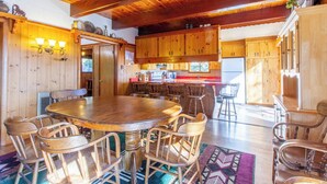 Un interior de madera con una mesa redonda rodeada de sillas, una zona de cocina con armarios de madera, y un ventanal que proporciona luz natural.