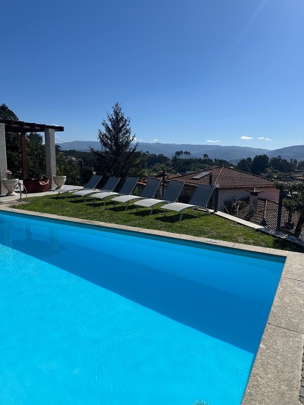 Piscina con sillas de descanso, una pérgola y vistas a montañas distantes.