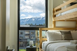 A bedroom with a bunk bed, a view of mountains through a large window, and a small table with a lamp. - Rest & Roam in Mackenzie Village (Revelstoke)