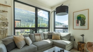 Ein modernes Wohnzimmer mit einer Eckcouch, großen Fenstern, Blick auf Berge, einem Holztisch und einer Stehlampe.