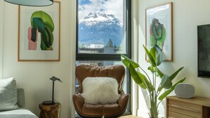 Ein Zimmer mit braunem Ledersessel, einem weißen, flauschigen Kissen, einem großen Fenster mit Blick auf die Berge und zwei gerahmten Kunstwerken.