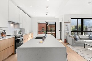 Une cuisine moderne avec un îlot central, un coin repas doté d’un canapé gris et une vue sur un immeuble grâce à de grandes fenêtres.