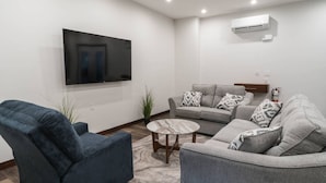 Een woonkamer met een flatscreen televisie, grijze banken, een blauwe fauteuil en een kleine ronde tafel.