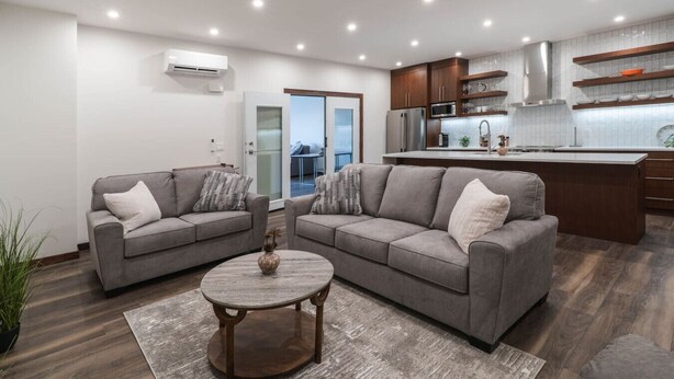 A modern living room with two grey sofas, a round coffee table, and a kitchen area with wooden cabinets and a stainless steel refrigerator. - The Alpine Den with Indoor Hot Tub (Revelstoke)