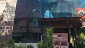 Een modern hotel met een glazen gevel die palmbomen weerspiegelt, een bord met daarop AVH Hotels & Restaurant, en potplanten ervoor.