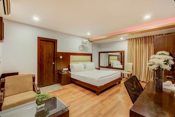 A hotel room with a bed, a sofa, a desk, a chair, a mirror, and a vase with flowers. - VH Hotels Mysore Road (Bengaluru)