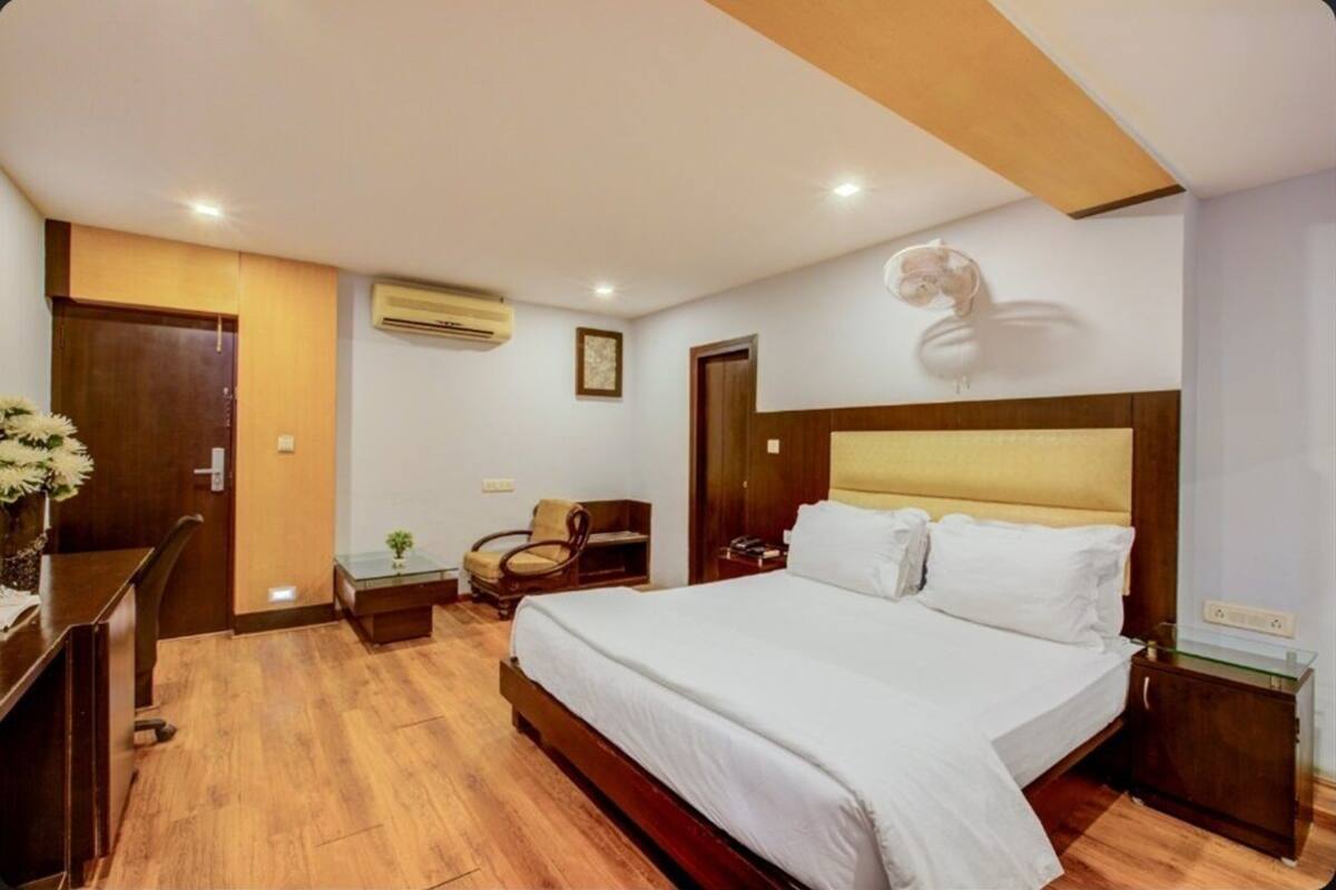 Habitación de hotel con piso de madera, cama con ropa blanca, escritorio con silla, una mesita con planta y aire acondicionado de pared.