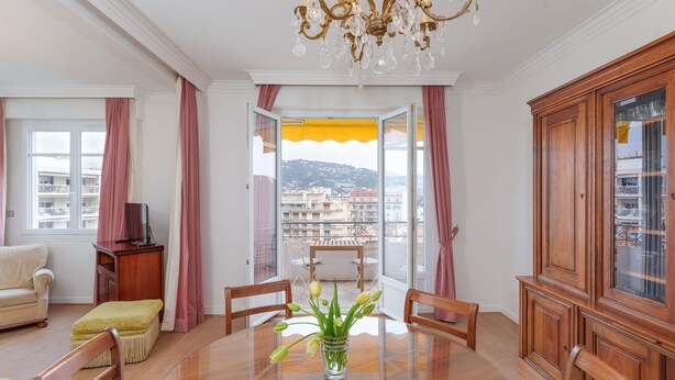 Ein Zimmer mit Esstisch, Stühlen, einer Chandelierleuchtre, einer Couch, einem TV-Schrank und Blick auf einen Balkon mit gelber Markise.