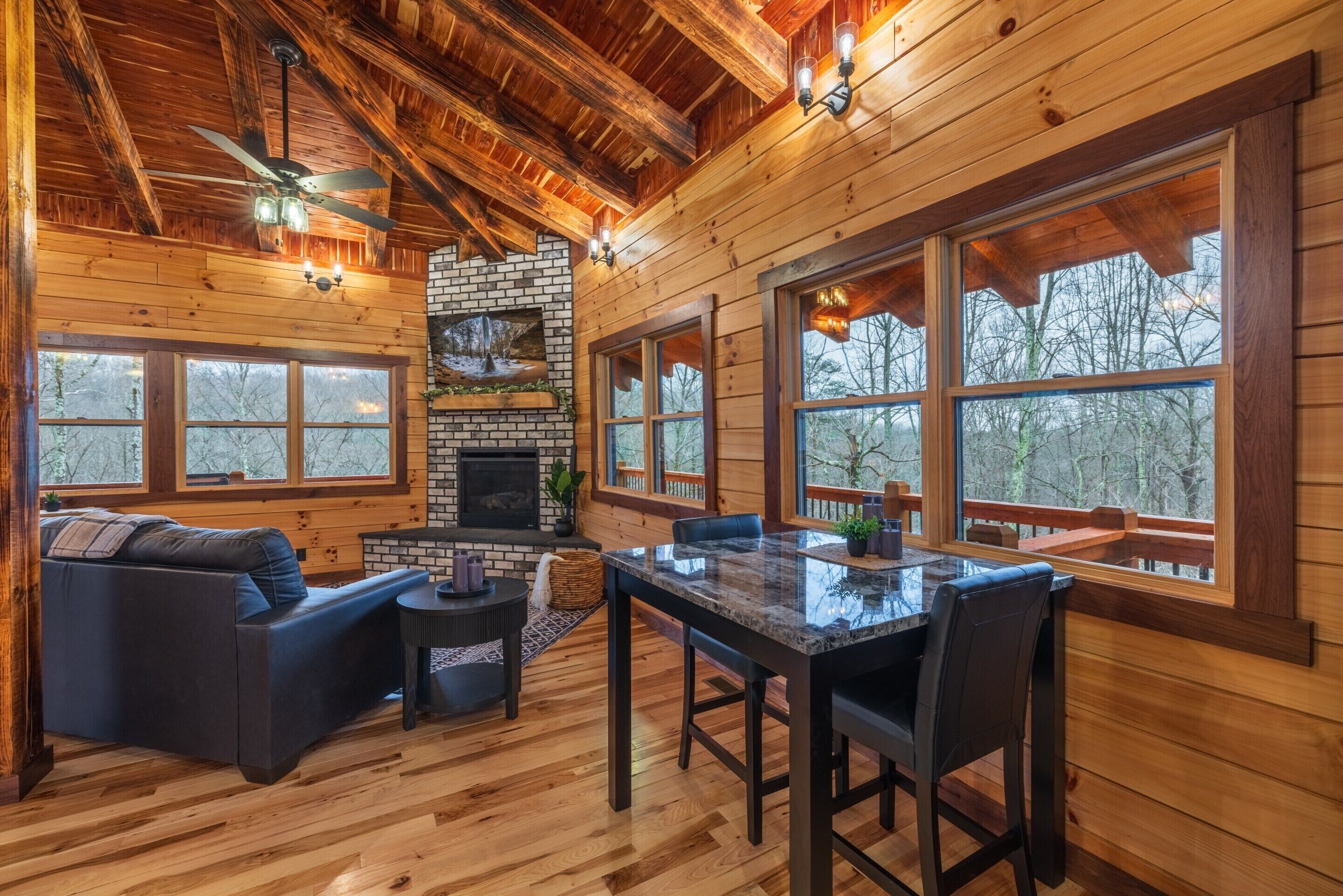 A log cabin interior with a fireplace, wooden walls, large windows, a dining area, and a ceiling fan.