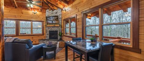 A log cabin interior with a fireplace, wooden walls, large windows, a dining area, and a ceiling fan.
