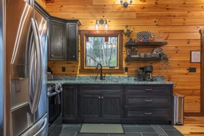 Eine moderne Küche mit dunklen Holzschränken, einem Edelstahlkühlschrank, einer Granitarbeitsplatte, einem Fenster mit Blick auf Bäume und einem kleinen Regal mit Dekoration.