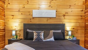 Ein Zimmer mit Holzwänden, einem Bett, Kissen, einem Nachttisch mit Lampe und einem gerahmten Zitat an der Wand.