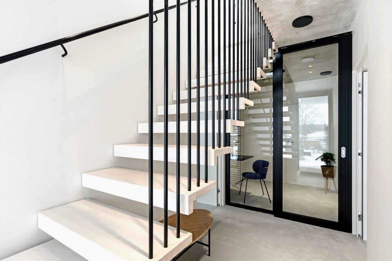 Eine moderne Treppe mit weißen Stufen und schwarzen Geländern, die zu einem Raum mit Glastür und Außenblick führt.