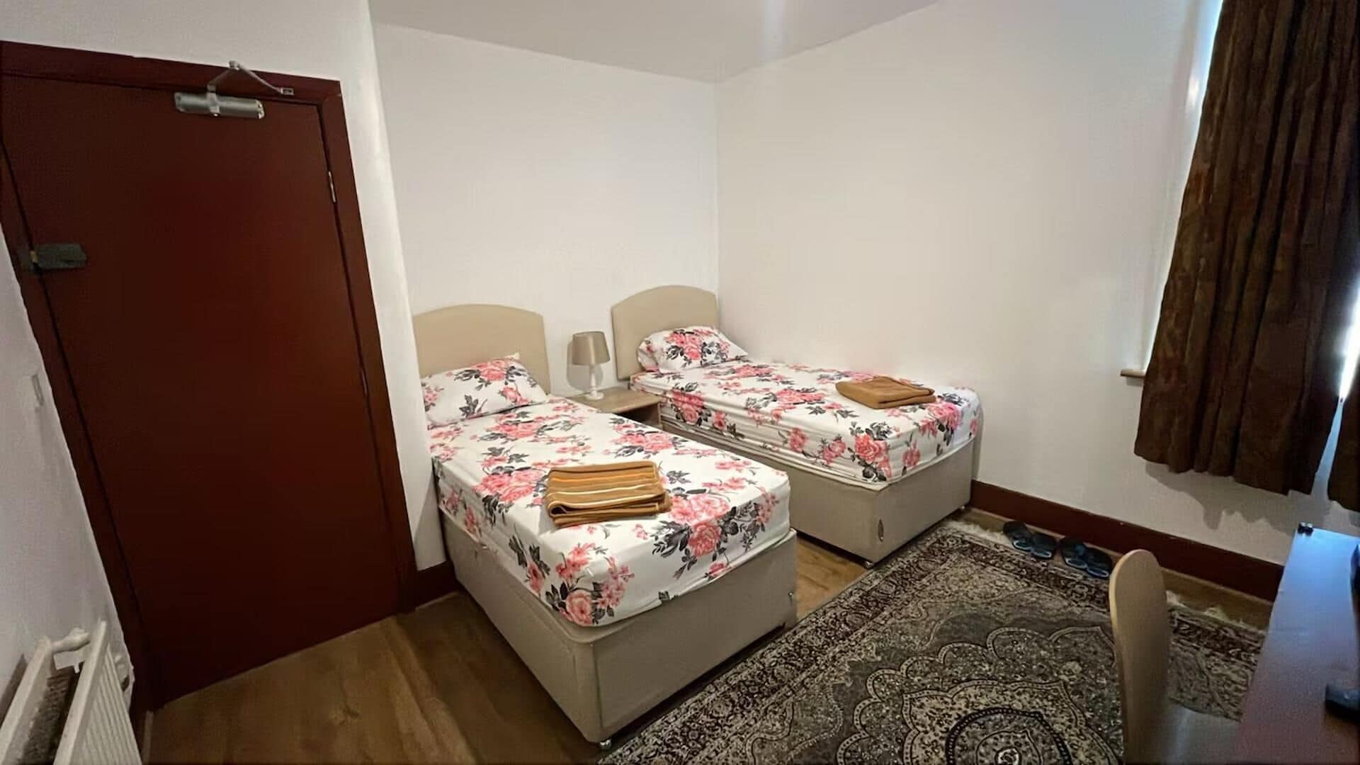 Una habitación pequeña y limpia con dos camas, una puerta, un ventanal con cortinas y una alfombra.