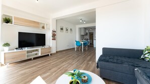 Ein modernes Wohnzimmer mit Flachbildfernseher, Holzboden, einem grauen Sofa, einem Essbereich mit blauen Stühlen und einer Küche im Hintergrund.