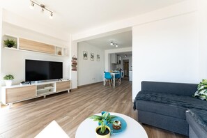 A modern living room with a flat-screen TV, wooden flooring, a grey sofa, a dining area with blue chairs, and a kitchen in the background. - Apartment in the Historic Centre of Malaga (Málaga)