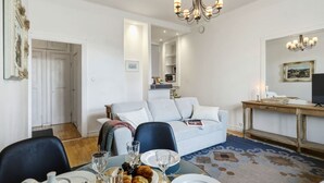 A dining area with a table set for two, a sofa, a chandelier, and a television on a wooden stand.
