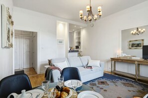A dining area with a table set for two, a sofa, a chandelier, and a television on a wooden stand. - Design District | Central & Quiet | 200m tram/bus (Helsinki)