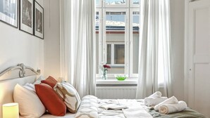 A hotel room with a bed, a window with curtains, a view of buildings, and a vase with flowers on the windowsill.