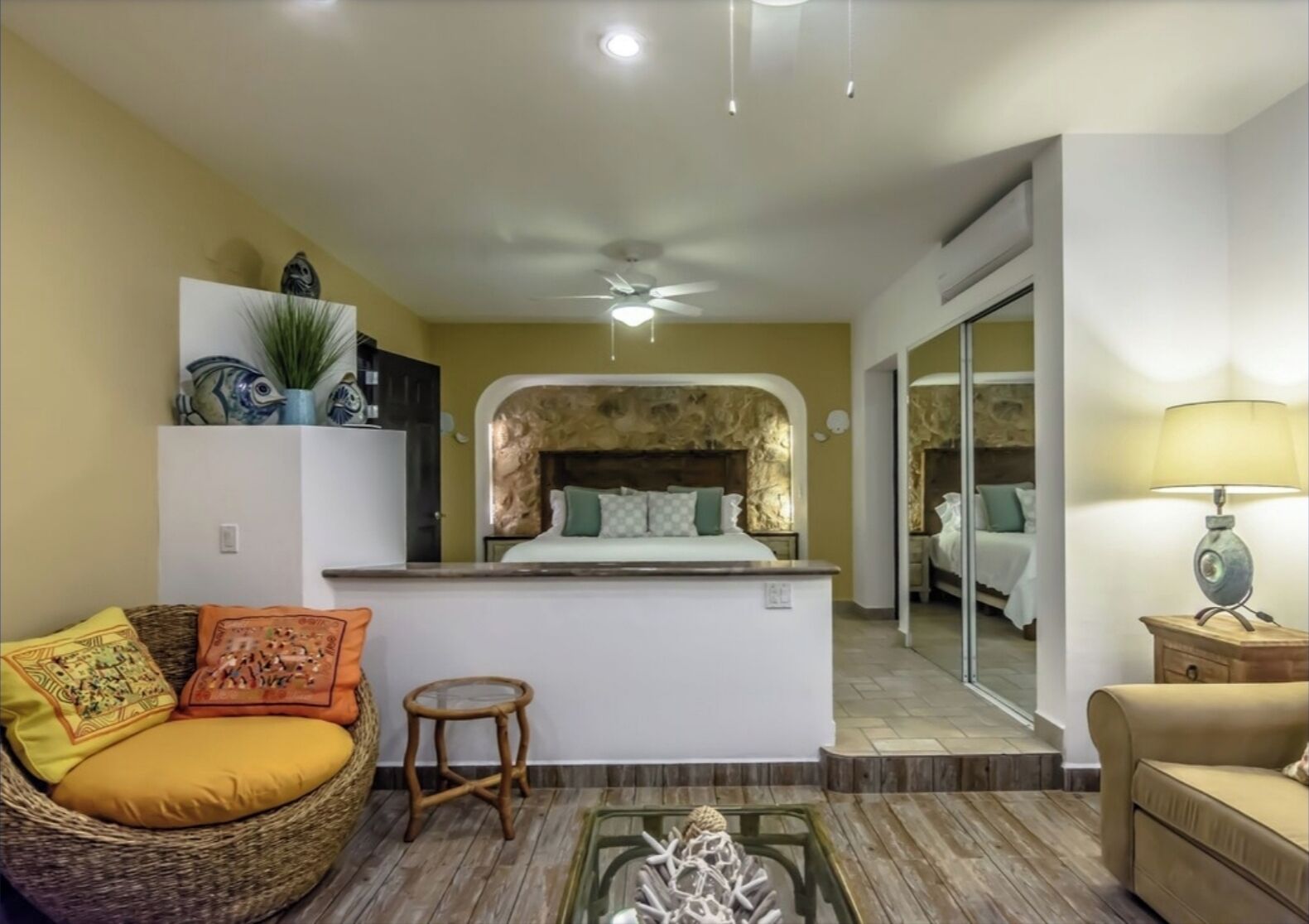 A compact hotel room with a bed, a small sitting area, a kitchenette, and a bathroom visible through a glass door.