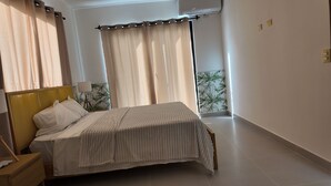 A bedroom with a bed, bedside table, lamp, curtains, and a wall-mounted air conditioner. - Elegant 3-bedroom Villa in Harmony Village Vista Cana (Punta Cana)