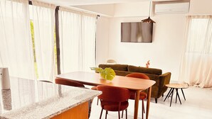 A modern living room with a dining area, a sofa, a TV mounted on the wall, and a window with curtains. - Elegant 3-bedroom Villa in Harmony Village Vista Cana (Punta Cana)