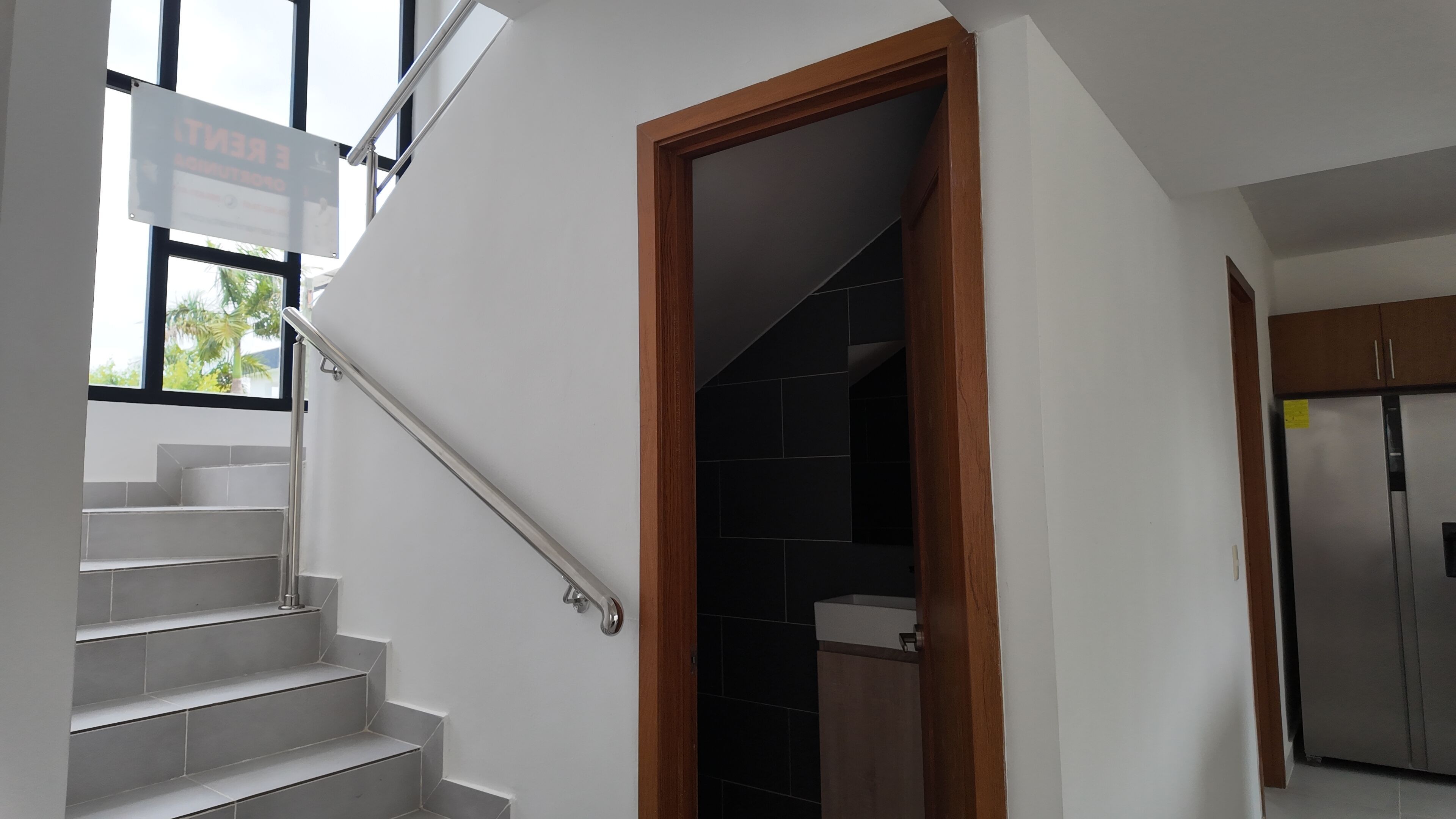 Un escalier avec une rampe, une porte en bois donnant sur une pièce sombre carrelée, et un coin cuisine avec un réfrigérateur.