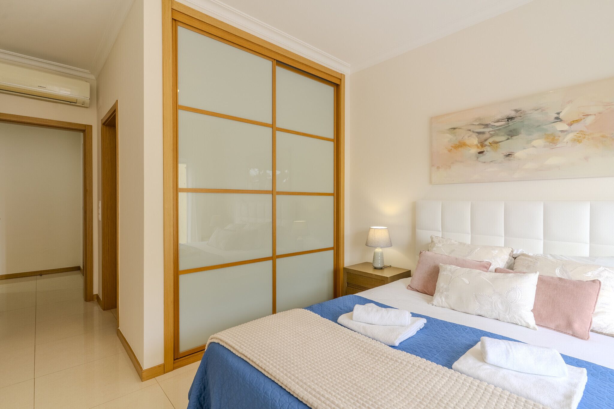 A hotel room with a bed, a nightstand, a lamp, a large window with blinds, and a painting on the wall.