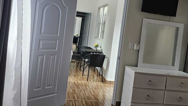 Een open deur die leidt naar een eetgedeelte met een tafel en stoelen, een dressoir met lades en een wandtelevisie.