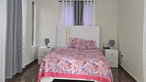 Een slaapkamer met een bed, een plafondventilator, een raam met gordijnen, een nachtkastje met een lamp en een airconditioning.