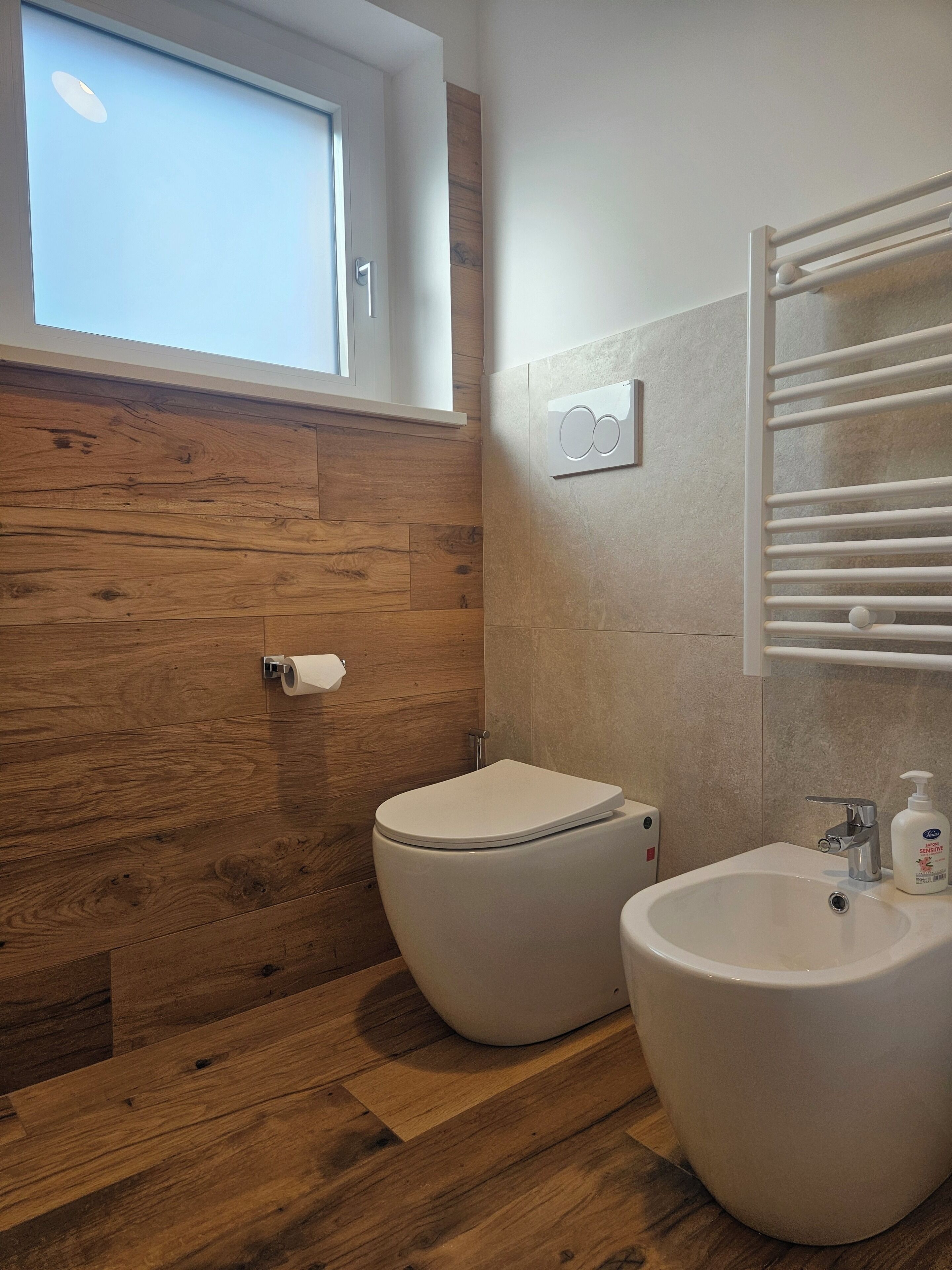 A bathroom with a toilet, bidet, and sink, wooden flooring, a window, and a towel rack.