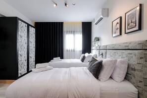 Quarto de hotel com duas camas, um guarda-roupa, uma janela grande com cortinas e quadros nas paredes.